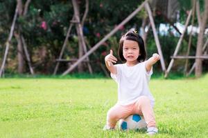 Portrait image of 4 baby kid. Happy Asian child girl sitting on white-blue football. Children gave a thumbs up showing that it was great. Learning sport and playing concept. Smiling little toddler. photo