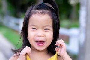 Close-up of happy children Asian are laughing and smiling sweetly with a cheerful cheerfulness. The kid girl is in a good mood, using two fingers to pull on the yellow shirt. Cute child 3 years old. photo