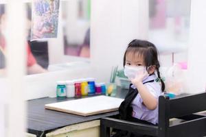 concept of taking care of oneself when doing external activities. Asian schoolgirl is going to learn to paint on a white canvas. Child turned to look at the camera and waved. Cute kids wearing mask. photo