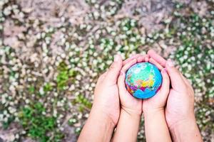 manos de niños y adultos abrazan el mundo. fondo de hierba con pequeñas flores blancas borrosas. concepto de preservar el mundo para la próxima generación. foto