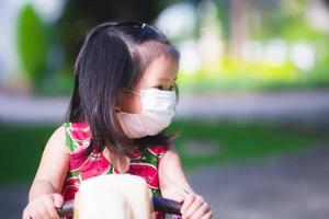 Girl play with toys rocking horse in playground. Children wear white medical face masks against PM2.5 dust and coronavirus disease Covid-19 outbreak. Little kid wearing red dress is 3 years old. photo