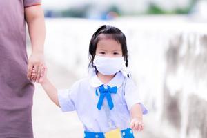 una colegiala asiática camina de la mano con una madre o un tutor. ir a la escuela. los niños usan uniformes escolares y mascarillas de tela para prevenir infecciones por virus o polvo, humos tóxicos de la contaminación del aire foto