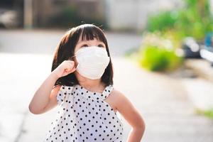 una chica adorable con una mascarilla médica sale a caminar afuera. niña feliz miró hacia arriba y estaba cuestionando algo con lo que se había encontrado. niño de 4-5 años vestir de blanco - tela negra estampado de lunares foto