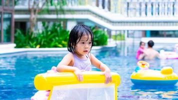 Asian child in the pool. Kid holding yellow rubber raft. Girl wearing purple swimming suit. Children with wet hair and body from playing in the water. Summer time. Baby aged 4-5 years old. photo