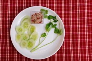 Yum small tuna salad in a clear plastic cup and place it on a white plate with coriander and sliced cucumber. Red plaid background. Top view. photo