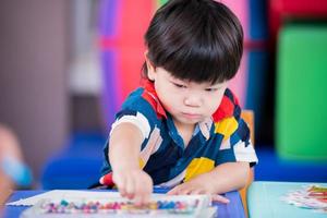 adorable niño asiático alcanzó a recoger pintura de tiza, bebé hizo arte en el papel colocado sobre la mesa. el niño aprende manualidades en casa. un pasatiempo para niños pequeños, 2-3 años. hijo estaba vestido con colores brillantes. foto