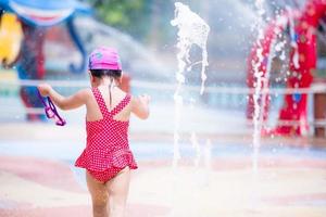 vista trasera de una niña pequeña en el parque acuático. niños con traje de baño rojo. niño con gafas de natación moradas y corriendo o bailando para jugar en el patio de la fuente. en el verano. foto