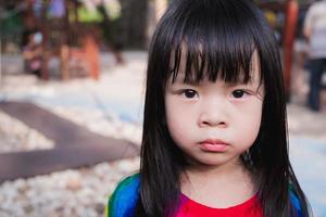 Closeup face child is hot sweat. Upset girl in the playground outside. Kid black haired unhappy when hot weather. Baby wear colorful dress aged 4 years old. In summer or spring time. photo