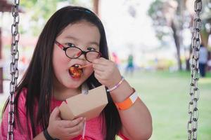 Portrait image of kid 5-6 years old. Cute Asian girl eating snacks from paper plate. Child sit on swing. Children wear eyeglasses due to myopia. Summer activities. Empty space for entering text. photo