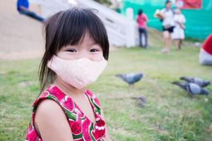 Cute Asian girl wearing mask sitting on grass. Behind the little child was flock of pigeons on green lawn. Blur people walking in park in evening. New Normal. Prevent spread of virus and dust PM2.5. photo