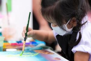 Pupils girl are concentrating on drawing and painting with brush and watercolor on the canvas. An Asian little child is wearing a white cloth face mask while learning to paint. Child 3 years old. photo