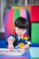 Portrait vertical image of baby boy picking up chalk colors for crafting on the table. Son doing art on white paper. Happy child in the hobby time. Homeschool. Homework of preschooler. Toddler cute. photo