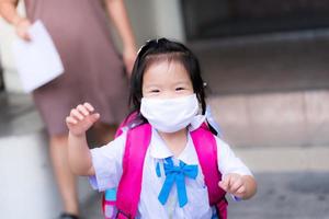 Mother or guardian takes the child back from school. A preschooler ran to see his father carrying a camera with happiness and a smile under a white cloth mask. 3-year-old girl carrying a pink backpack photo