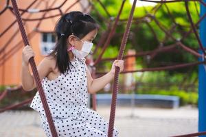 Portrait image child 5-6 years old. Children having fun climbing red rope netting. Little girl wear white face mask to prevent small dust particles pm2.5 and spread virus. Kid play playground outdoors photo