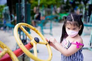 Portrait image kid 5-6 years old. Cute girl wears mask while playing exercise equipment in park. Children are learning interestingly with playground equipment. Child try to turn yellow garlands. photo