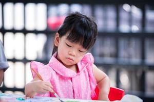 Asian kid doing art with water color brush on desk at home. Cute girl happy learning craft. Child aged 4 years old, wearing pink t-shirt. photo