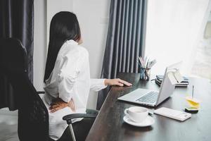 Young asian woman is sitting at work and has tension pain in her neck. Concept office syndrome backache pain from occupational disease, Female having muscle pain from using laptop computer, back pain. photo