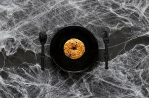 Scary halloween dinner party concept with funny donut face on black plate, knife and fork with spider and cobweb on black background. photo