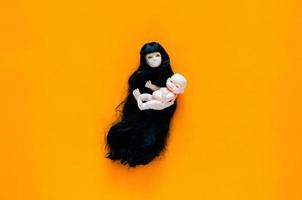 Long hair flying female ghost doll carries her baby on orange background. Minimal Halloween scary concept. photo