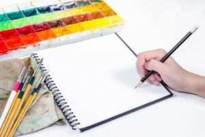 mano humana sosteniendo un lápiz con un cuaderno y una bandeja de colores foto