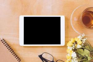 Top view of tablet with tea cup, flower, notebook and glasses on wooden table photo