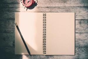 Top view of notebook with pencil and clock on wooden table background. Vintage tone photo