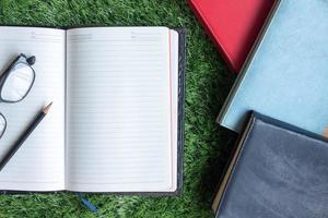 Blank notebook with pencil, glasses and books on green grass background. photo