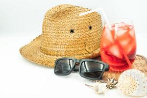 sombrero con gafas de sol, cóctel rojo y conchas marinas sobre fondo blanco, concepto de vacaciones de verano foto