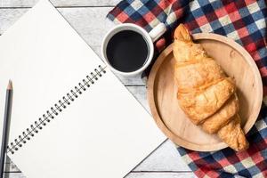 Top view of blank notebook with pencil, croissant and coffee on wooden table background. photo