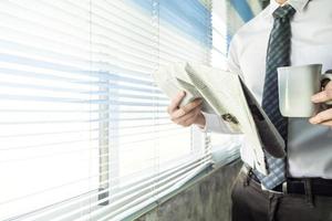 Businessman reading newspaper and holding coffee cup while standing at a window in an office. photo