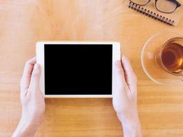 Top view of hands holding tablet, tea cup with notebook and glasses on wooden table photo