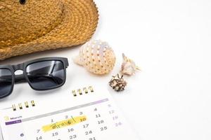 sombrero con gafas de sol, calendario y conchas marinas sobre fondo blanco, concepto de vacaciones de verano foto