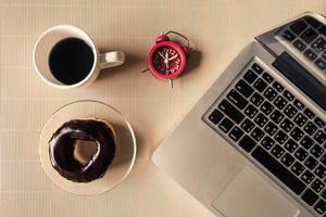 Top view of laptop with coffee cup, donut and clock on table. photo