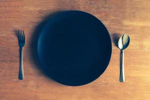 Top view of empty black plate with spoon and fork on wooden table