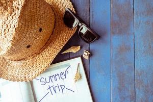 sombrero con gafas de sol, calendario y conchas marinas sobre fondo de madera pintada de azul. concepto de vacaciones de verano foto