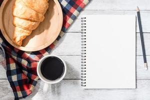 Top view of blank notebook with pencil, croissant and coffee on wooden table background. photo