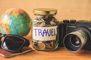 Coins in glass jar with accessories of traveler on wooden table. Saving money for travel concept photo