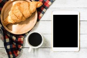 Top view of tablet with croissant and coffee on wooden table background. photo