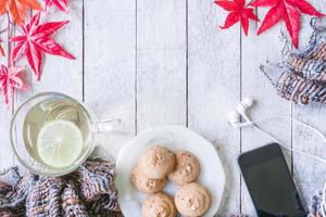 taza de té con limón, galleta, bufanda, auriculares, teléfono inteligente y hojas de arce en la mesa de madera. foto