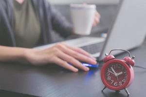 primer plano del reloj rojo en el escritorio, hora de la tarde, mujer joven casual usando una computadora portátil foto