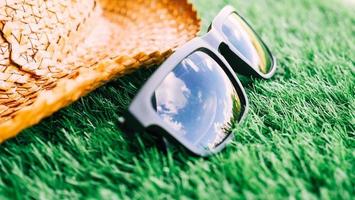 Sunglasses with the reflection of  blue sky and hat on green grass, Soft focus. photo