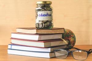 Coins in glass jar with education label, books,glasses and globe on wooden table. Financial concept. photo