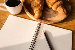 Top view of blank notebook with pencil, croissant and coffee on wooden table background. photo