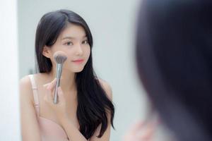 Beauty portrait young asian woman smiling with face looking mirror applying make up with blusher cheek in the room, happy beautiful girl blush applying facial, skin care and cosmetic concept. photo