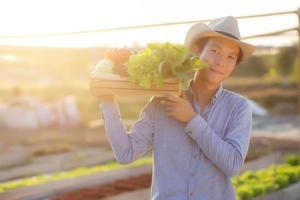 Portrait young asian man smiling harvest and picking up fresh organic vegetable garden in basket in the hydroponic farm, agriculture and cultivation for healthy food and business concept. photo