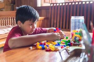 Asian boy is getting creative with assembling colorful plastic bricks into robots and planes on a wooden table happy and fun at home.Kid Creators concept. photo