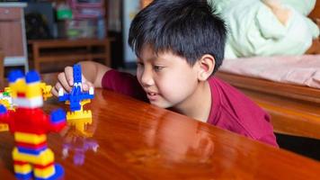 Asian boy is getting creative with assembling colorful plastic bricks into robots and planes on a wooden table happy and fun at home.Kid Creators concept. photo