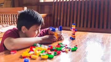 Asian boy is getting creative with assembling colorful plastic bricks into robots and planes on a wooden table happy and fun at home.Kid Creators concept. photo