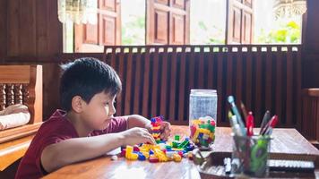 el niño asiático se está volviendo creativo al ensamblar coloridos ladrillos de plástico en robots y aviones en una mesa de madera feliz y divertido en casa. concepto de creadores de niños. foto