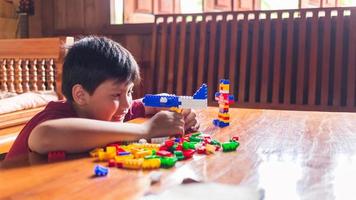 Asian boy is getting creative with assembling colorful plastic bricks into robots and planes on a wooden table happy and fun at home.Kid Creators concept. photo
